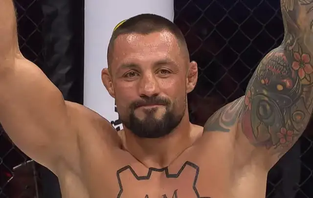 Mateusz Janur, wojownik MMA, z podniesionymi rękami i tatuażami na ramionach i klatce piersiowej, świętuje zwycięstwo.