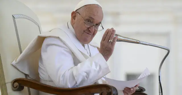 Papież Franciszek w białej szacie siedzi na krześle, trzymając mikrofon i kartkę.