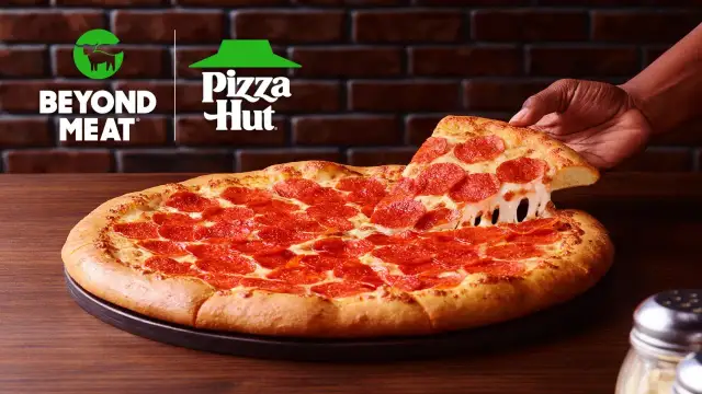 Pizza pepperoni z ciągnącym się serem, podnoszona przez rękę. Logo Beyond Meat i Pizza Hut w tle.