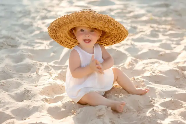 Uśmiechnięte dziecko w słomkowym kapeluszu na plaży. Idealne miejsce, gdzie na wakacje z półrocznym dzieckiem, by cieszyć się słońcem i piaskiem.