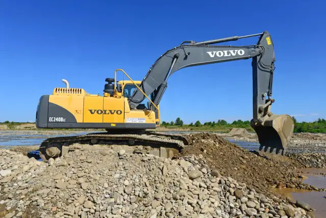 Żółta koparka Volvo EC240B LC pracuje na kamienistym brzegu rzeki. Zastanawiasz się, ile kosztuje godzina pracy koparki?