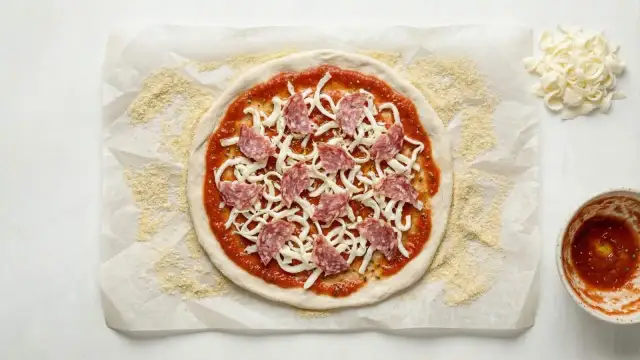 Okrągłe ciasto na pizzę z sosem, serem i salami, przygotowane na papierze do pieczenia.