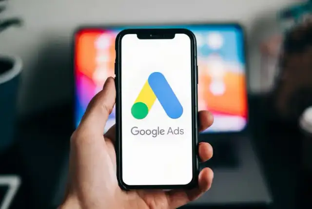 Dłoń trzyma smartfon z logo Google Ads. W tle widać ekran laptopa. Dziś budżet w reklamie Google Ads ustalamy.