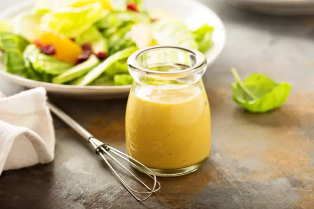 Ein Glas mit gelbem Blattsalat Dressing steht neben einem Schneebesen. Im Hintergrund ein Teller mit frischem Salat.