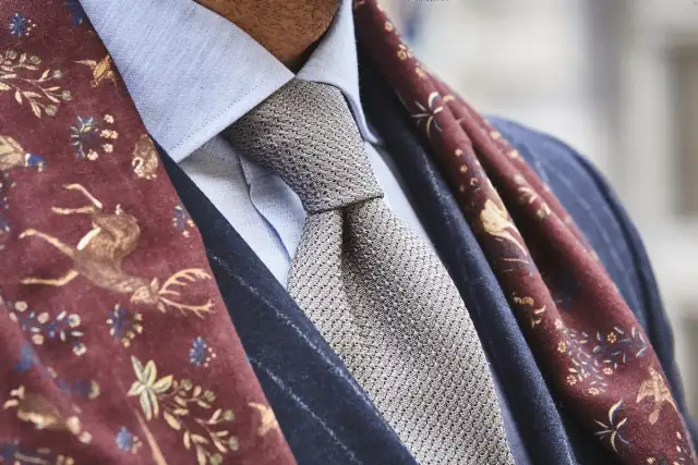 Elegancki mężczyzna w błękitnej koszuli, szarym krawacie i bordowym szalu z motywem jeleni. Całość emanuje nonszalancką elegancją, czyli sprezzaturą.