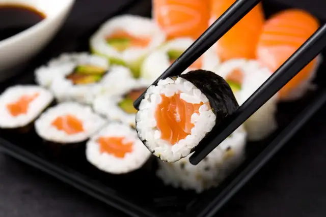 Pałeczki trzymające kawałek sushi z łososiem na tle talerza z różnymi rodzajami sushi.