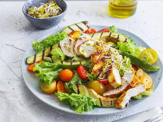 Gegrillter Hähnchensalat mit Avocado, Tomaten und Sprossen auf einem Teller.