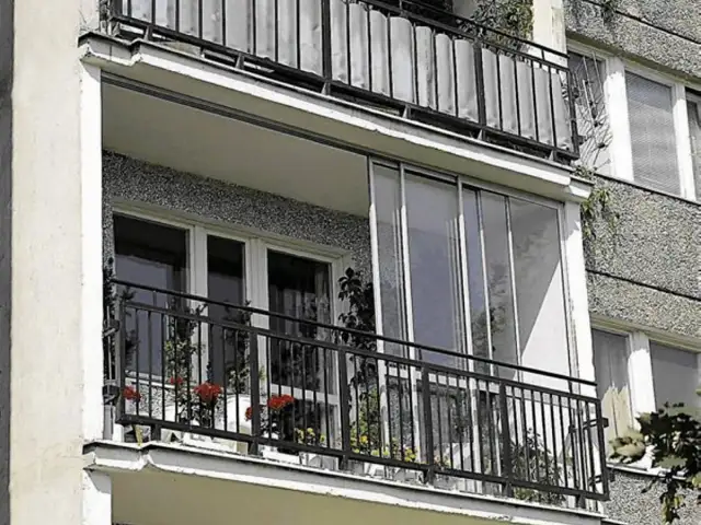 Zabudowa balkonu w bloku z przeszklonymi drzwiami i metalową balustradą. Na balkonie kwiaty w doniczkach.
