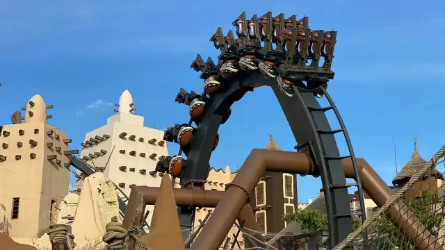 A roller coaster with brown seats and patterned spheres against a blue sky and themed buildings.