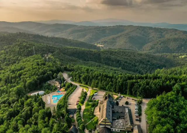 Widok z lotu ptaka na hotel SPA z basenem i zjeżdżalnią, otoczony lasem i górami.