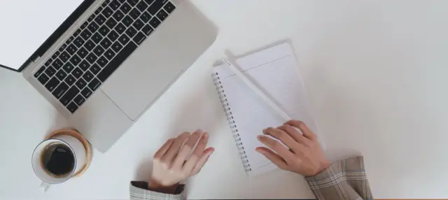 Ręce piszą CV po niemiecku przy laptopie, notatniku i kawie.