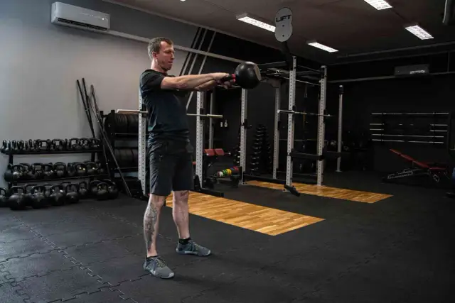 Mężczyzna ćwiczy z kettlebell w siłowni. W tle stojaki z ciężarami i sprzęt do treningu siłowego.