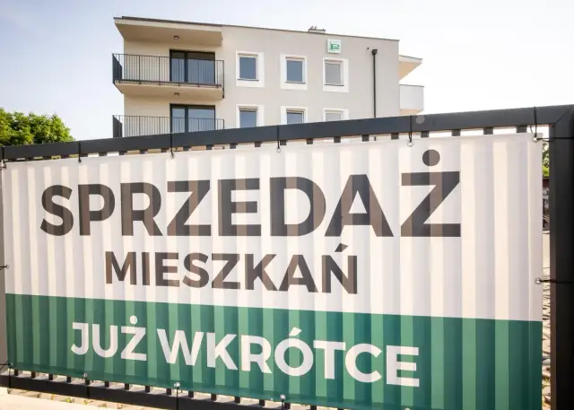 Baner z napisem "SPRZEDAŻ MIESZKAŃ JUŻ WKRÓTCE" na tle nowoczesnego budynku mieszkalnego.