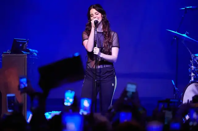 Lana Del Rey, w czarnej, prześwitującej bluzce i sportowych spodniach, śpiewa na scenie. Fani z telefonami tworzą tłum. To jej lana del rey opener.