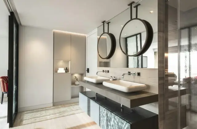 Un moderno baño con dos lavabos sobre encimera de piedra, espejos circulares colgantes y armarios integrados. Se puede hacer un baño en una habitación así.