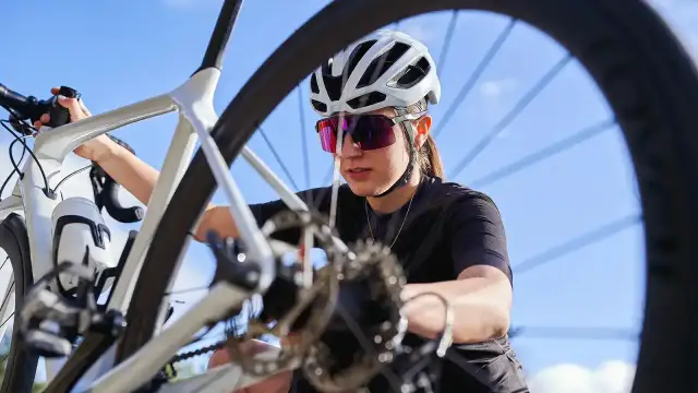 Kobieta w kasku i okularach rowerowych sprawdza rower. Widoczny jest fragment ramy, kierownicy i koła, a także przerzutka.