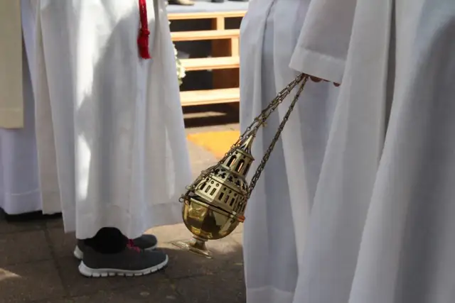 Osoba w białej szacie trzyma złote kadzidło na łańcuszku.