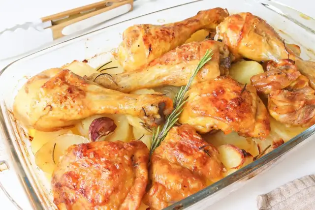 Muslos de pollo al horno con patatas, dorados y jugosos, acompañados de patatas asadas, ajo y romero en una fuente de cristal.