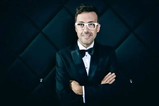 DJ Adamus w smokingu i białych okularach, z rękami skrzyżowanymi na piersi, na tle pikowanej czarnej ściany.