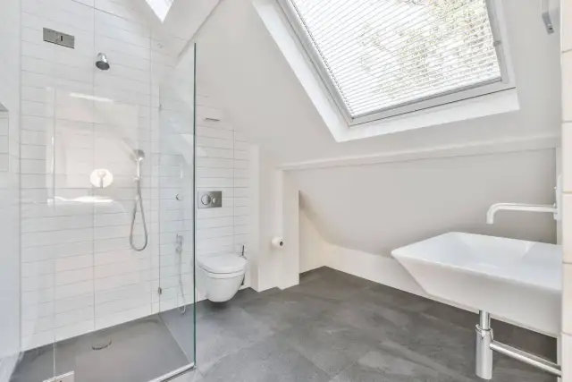 Moderno baño en buhardilla con ducha de cristal, inodoro y lavabo bajo una claraboya.