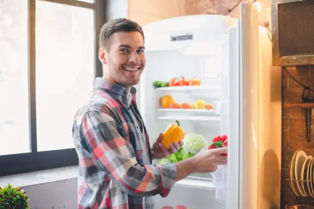 Hombre sonriente saca pimientos de la nevera. Se pregunta cuánto dura el pisto en la nevera, mientras organiza sus verduras.