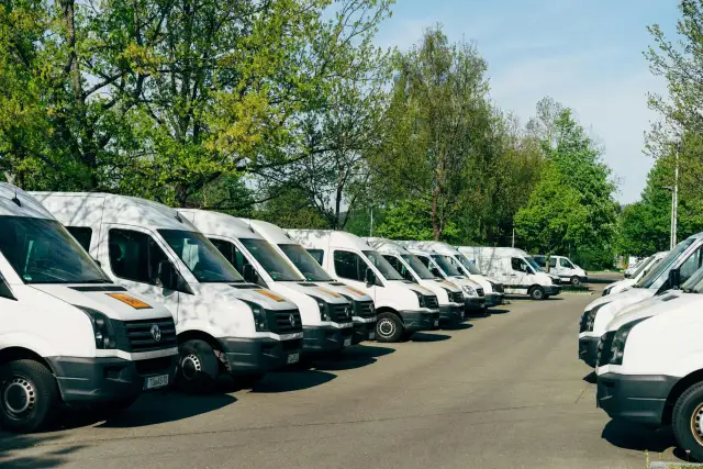 Rząd białych busów zaparkowanych pod drzewami. Zastanawiam się, jaki bus w benzynie sprawdziłby się najlepiej do przewozu towarów.