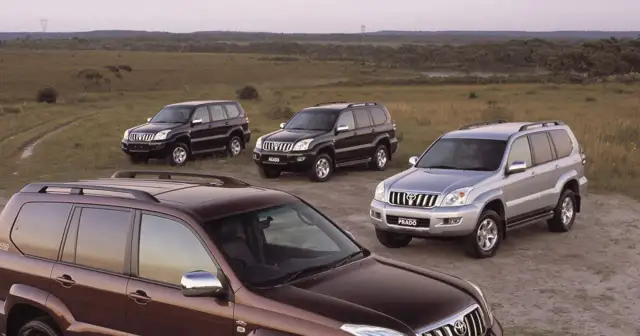 Cztery samochody Toyota Land Cruiser 120, w tym jeden brązowy, dwa czarne i jeden srebrny, stoją na polnej drodze wśród traw.