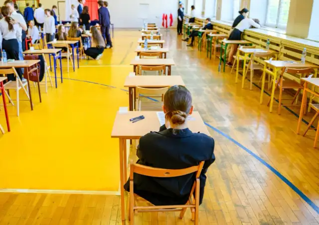 Uczniowie siedzą przy ławkach w sali gimnastycznej, przygotowując się do egzaminu dojrzałości.