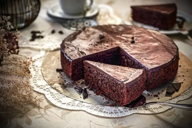 Ein saftiger Schokokuchen mit glänzender Glasur, ein Stück davon auf einem Tortenheber, daneben Schokoladenstücke und eine Kaffeetasse.