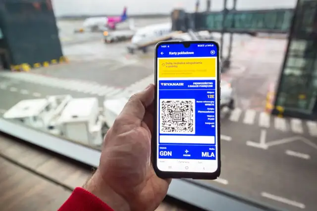 Dłoń trzyma smartfon z kartą pokładową Ryanair i kodem QR na tle lotniska.