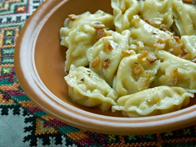 Pyszne pierogi ruskie z podsmażoną cebulką i skwarkami, podane w glinianej misce na haftowanej serwetce.