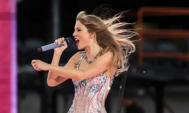 Taylor Swift w błyszczącej sukience śpiewa na koncercie w Warszawie, włosy rozwiane przez wiatr.