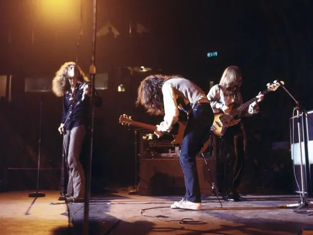 Energetyczny **Led Zeppelin koncert** na scenie. Wokalista z burzą włosów, gitarzysta pochylony nad instrumentem, basista grający w tle.