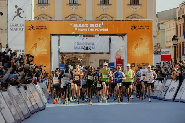 Biegacze startują w maratonie. Wielka brama z napisem "Cracovia Maraton" i hasłem "Masz moc!" symbolizuje wyzwanie, ile ma maraton.