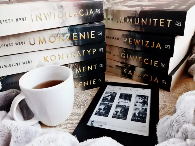 Stos książek Remigiusza Mroza, biały kubek z herbatą i czytnik Kindle.