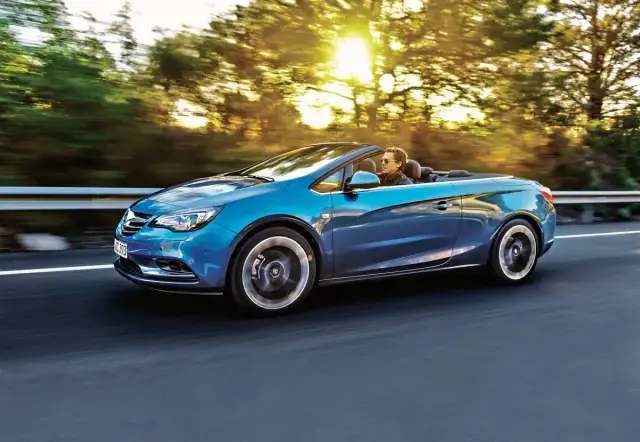 Niebieski Opel Cascada z otwartym dachem sunie po drodze, a słońce przebija się przez drzewa.