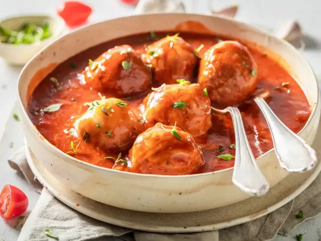 Albóndigas caseras con tomate frito, servidas en un cuenco de cerámica con hierbas frescas y dos cucharas listas para disfrutar.