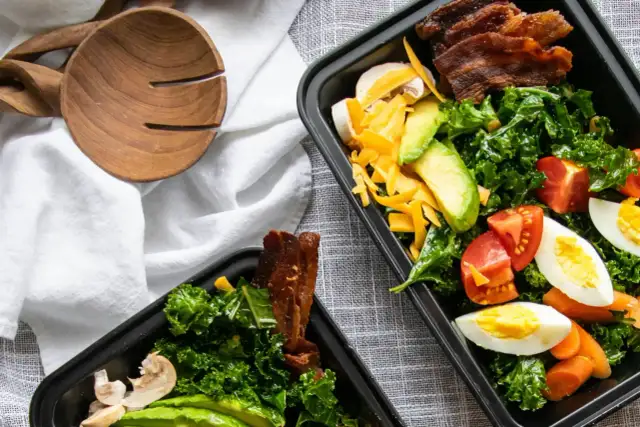 Zwei Behälter mit leckeren low carb meal prep Salaten, gefüllt mit Grünkohl, Avocado, Tomaten, Eiern, Käse und Speck.