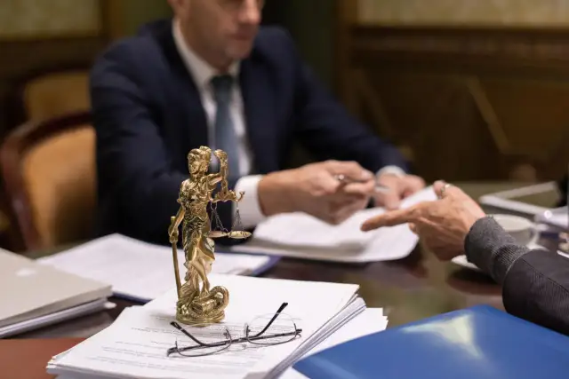 Figurka Temidy, dokumenty i okulary na stole, dwóch mężczyzn w garniturach przy biurku.