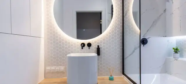 Moderno baño con lavabo blanco, espejo redondo iluminado y azulejos hexagonales. Ideal para tu presupuesto baño.