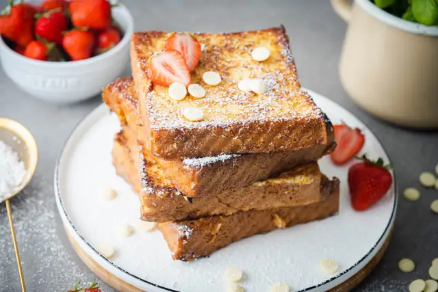 Gestapelte French Toast Scheiben, bestäubt mit Puderzucker und garniert mit Erdbeeren und weißen Schokoladenchips.