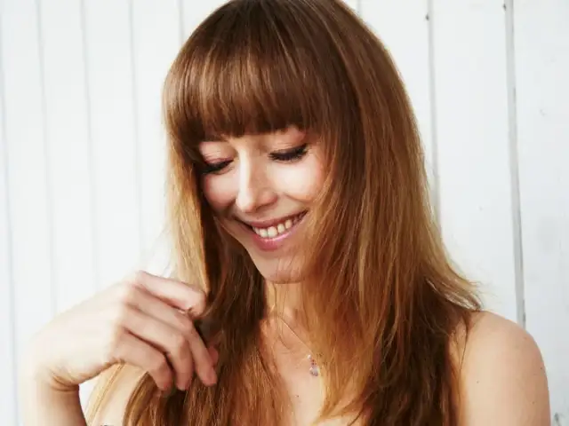 Uśmiechnięta kobieta z długimi, brązowymi włosami i grzywką, bawiąca się kosmykiem włosów.