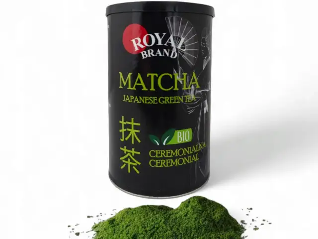 Czarna puszka z zieloną herbatą matcha Royal Brand i garść proszku matcha na białym tle.