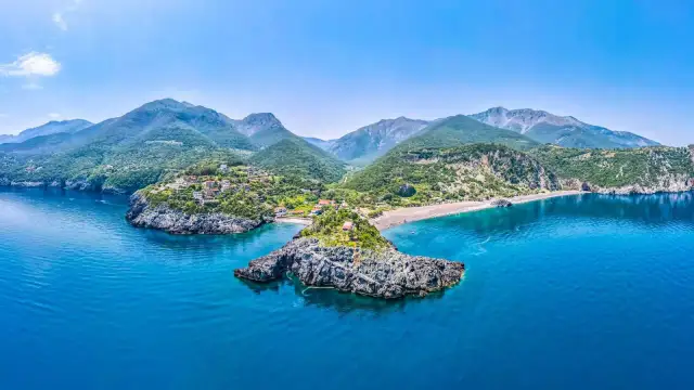 Idealne miejsce, gdzie na wakacje w sierpniu: malownicza zatoka z kamienistą wysepką, białe domki na zielonym wzgórzu i góry w tle.