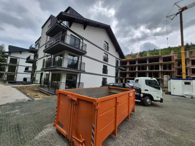 Pomarańczowy kontener na gruz przed nowym budynkiem i placem budowy.