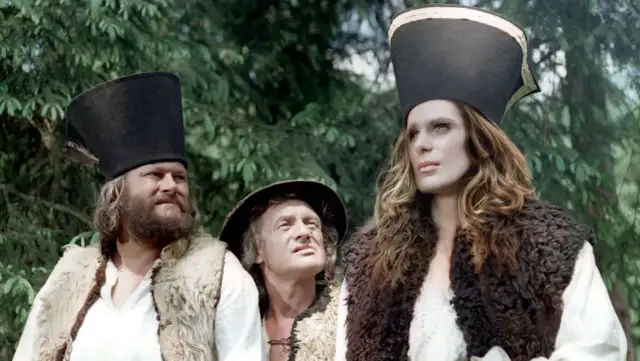 Trzej bohaterowie w strojach z baśni o 12 miesiącach: brodaty mężczyzna, starszy pan i młoda kobieta w futrzanych kamizelkach i wysokich czapkach.