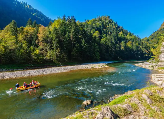 Żółta tratwa z ludźmi płynie po zielonej rzece w otoczeniu gór i lasów.