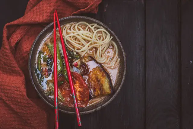 A miseczce z aromatycznym ramen wegetariańskim, pełnej makaronu, grillowanego bakłażana i pomidorów, spoczywają czerwone pałeczki.