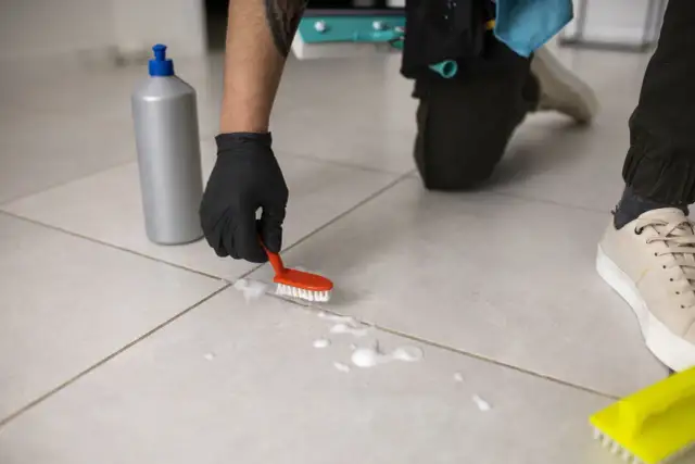 Osoba w czarnej rękawiczce czyści podłogę z płytek małą szczoteczką i pianą.
