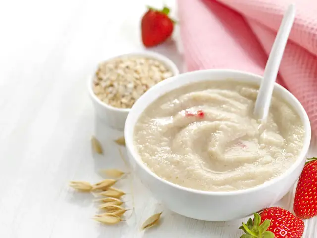 Owsianka z truskawkami, idealna do wprowadzania pokarmów u niemowląt. Na stole biała miseczka z kaszką, obok miseczka z płatkami owsianymi, świeże truskawki i różowy ręcznik.